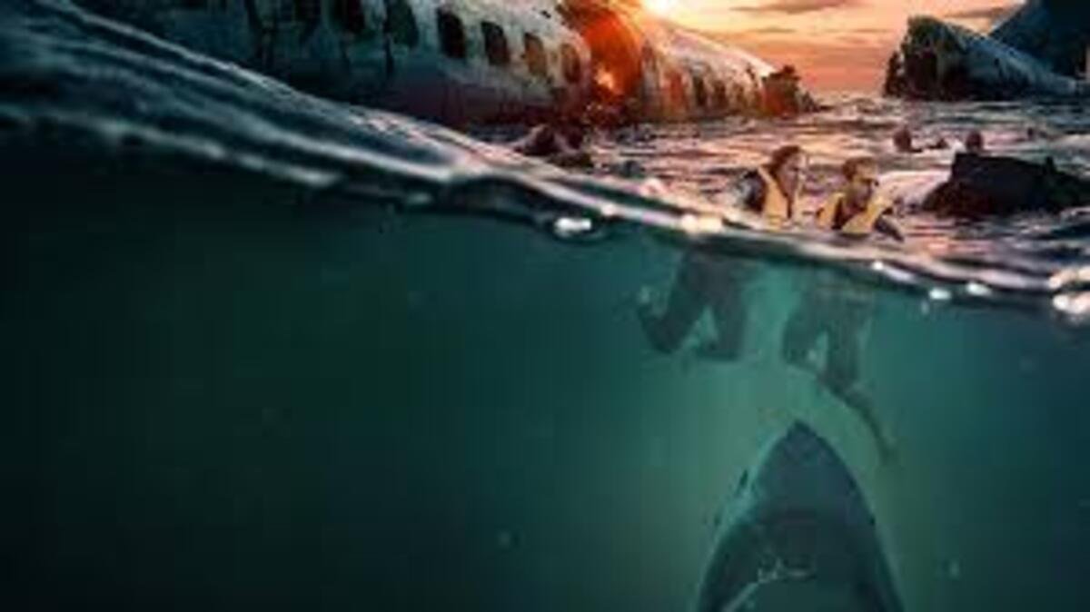 Composição visual dividida horizontalmente pela linha da água. Na metade superior, sobreviventes de um desastre aéreo flutuam no mar gelado e escuro ao entardecer, próximos aos destroços de uma fuselagem de avião que está parcialmente submersa. O céu ao fundo tem tons alaranjados de pôr do sol. Na metade inferior, abaixo da superfície da água escura e esverdeada, as pernas dos sobreviventes aparecem submersas enquanto a silhueta imensa e ameaçadora de um tubarão surge das profundezas, nadando em direção a eles.