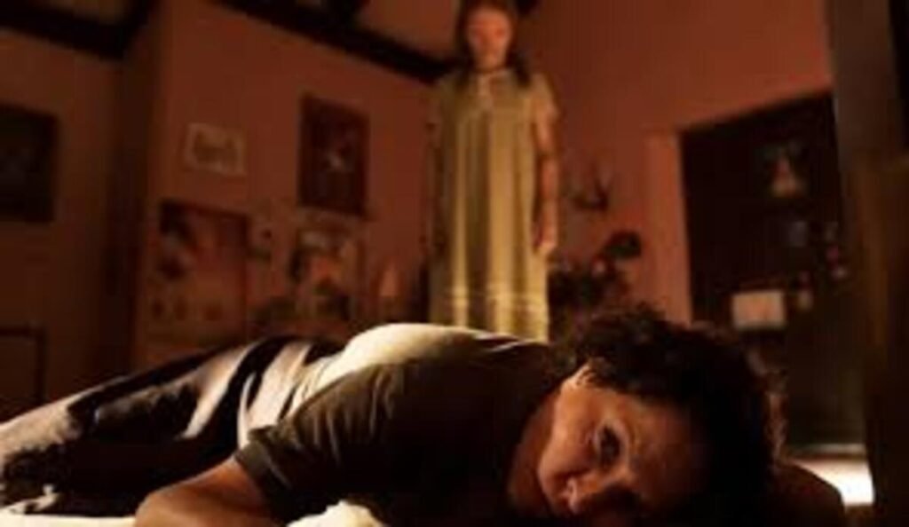 Crítica | Maldição da Múmia (2026): O terror visceral de Lee Cronin sobre luto e culpa 4 Cena de terror psicológico com iluminação sombria e avermelhada dentro de um quarto. Em primeiríssimo plano, uma mulher com cabelos escuros está caída de bruços no chão, com o rosto voltado para a tela, apresentando uma expressão de medo, cansaço ou desespero. Ao fundo, fora de foco e em pé no centro do quarto, uma figura feminina misteriosa vestindo uma camisola longa observa a situação de forma ameaçadora. O ambiente ao redor exibe quadros nas paredes e detalhes de uma residência antiga sob uma luz fraca.