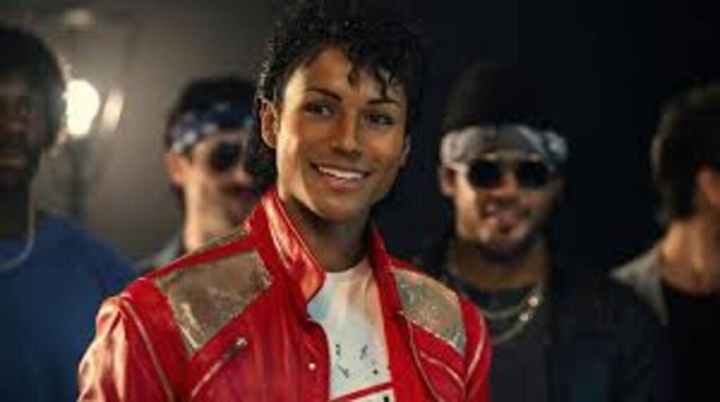 Primeiro plano do ator Jaafar Jackson sorrindo, caracterizado como Michael Jackson na era do álbum Thriller. Ele veste a icônica jaqueta de couro vermelha com detalhes prateados nos ombros. Ao fundo, outros atores aparecem desfocados, compondo o cenário de um set de filmagem ou videoclipe