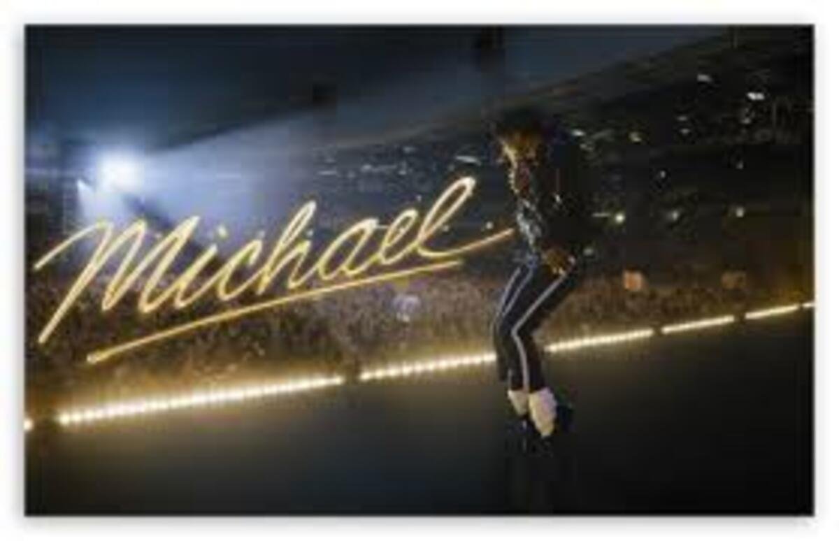 Fotografia em um palco escuro com luzes de estádio ao fundo, mostrando um artista em uma pose de dança icônica, inclinando-se para a frente. Sobreposta à imagem, está a assinatura estilizada 'Michael' em letras douradas e cursivas