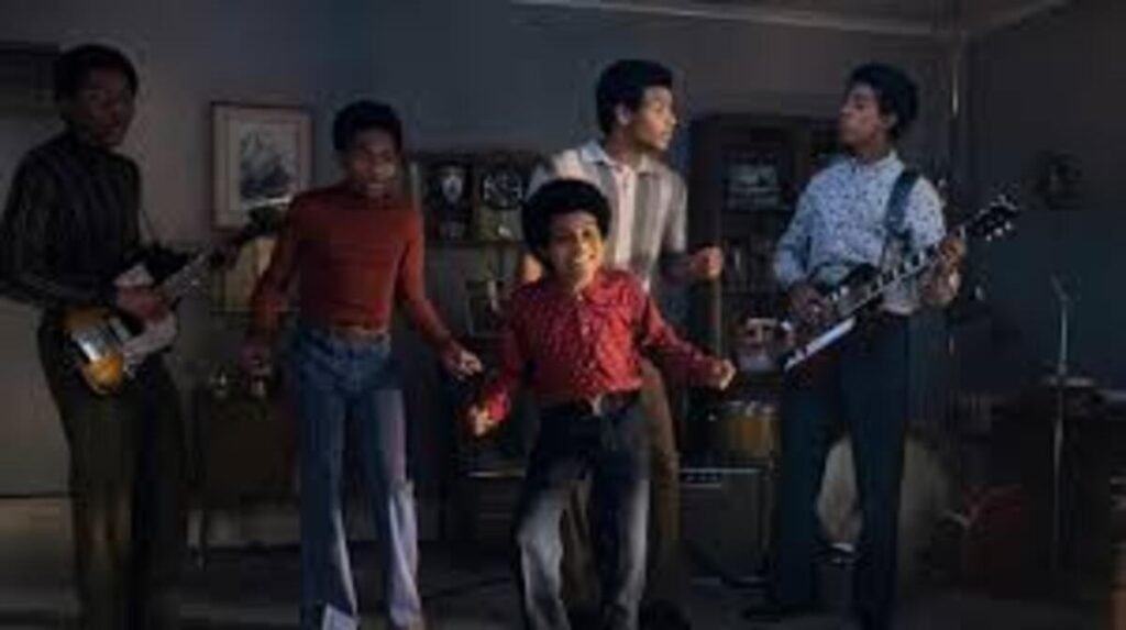 Cena do filme biográfico 'Michael' mostrando os atores que interpretam os Jackson 5 durante um ensaio na juventude. O grupo está reunido em uma sala, com o jovem Michael Jackson em destaque ao centro, sorrindo e cantando com seus irmãos