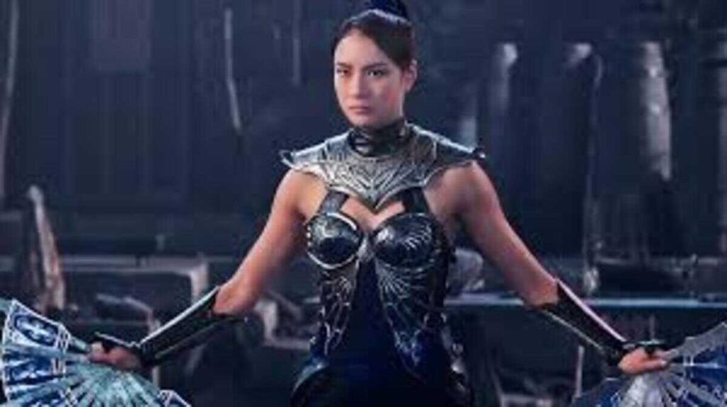 Primeiro plano da personagem Kitana, do filme Mortal Kombat, interpretada por uma atriz com expressão séria e cabelos presos em rabo de cavalo. Ela veste uma armadura tática preta e prateada detalhada. Kitana segura e exibe dois leques de metal prateado e azul, suas armas características, um em cada mão, com os braços ligeiramente estendidos para os lados. O fundo é um cenário industrial escuro e desfocado.