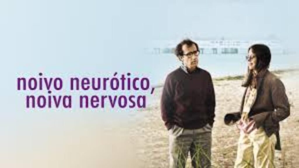 Cena do filme Noivo Neurótico, Noiva Nervosa em uma praia. À esquerda, o título do filme aparece em letras roxas minúsculas sobre um fundo claro e esfumaçado. Ao centro e à direita, os personagens interpretados por Woody Allen e Diane Keaton conversam de pé na areia clara. Ele veste um suéter escuro e calças cinzas, enquanto ela usa um casaco marrom e calças beges. Ao fundo, vê-se a linha do mar e uma estrutura que lembra um cais ou navio de forma sutil.