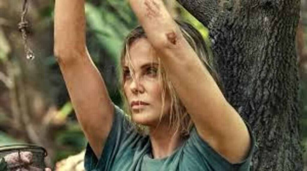 Uma mulher com cabelos loiros e úmidos, vestindo uma camiseta verde, aparece com os braços levantados. Ela tem uma pequena ferida visível no cotovelo e uma expressão de cansaço e determinação. Ao fundo, nota-se o tronco de uma árvore e vegetação, sugerindo um ambiente de floresta ou selva.