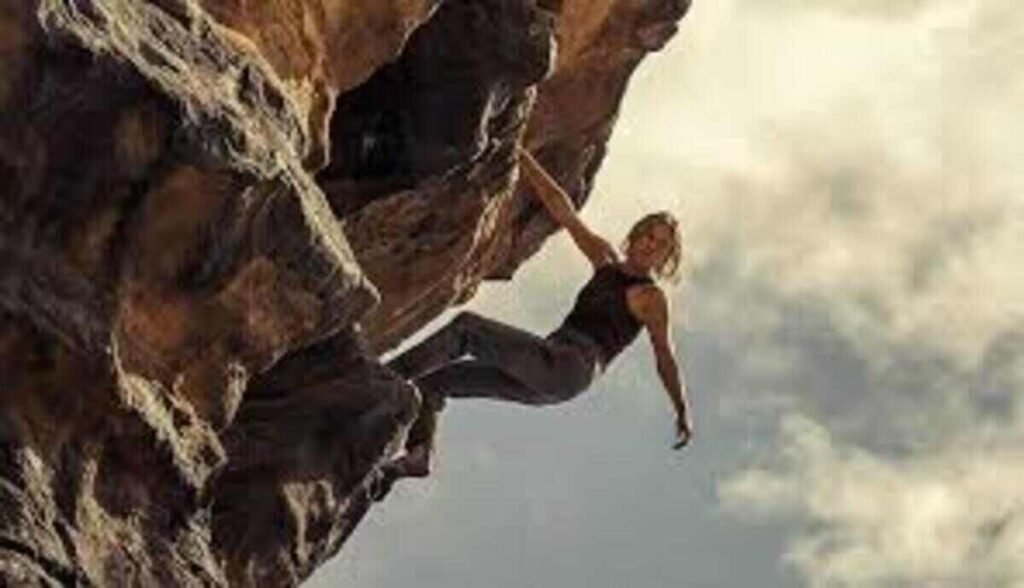 Uma mulher praticando escalada em uma formação rochosa íngreme. Ela usa uma regata preta e calças escuras, segurando-se com as mãos em uma saliência da pedra enquanto o corpo está suspenso sobre o vazio. Ao fundo, o céu está nublado com tons claros, destacando a altura e o perigo da cena.