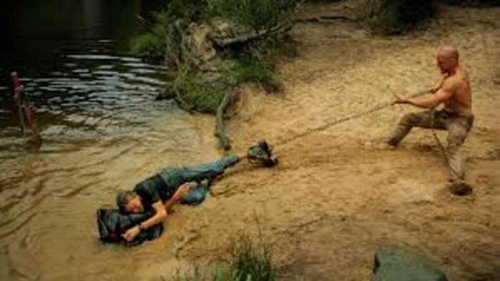 Cena de ação em uma margem lamacenta à beira de um rio. Um homem forte, sem camisa, está na areia puxando com esforço uma corda amarrada ao pé de uma mulher. A mulher está caída na água rasa, segurando uma mochila preta, enquanto tenta se segurar na margem. O cenário ao redor tem vegetação densa e raízes expostas.
