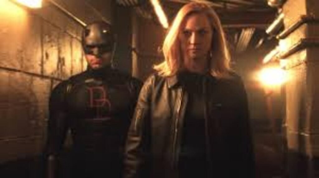 Karen Page e o Demolidor caminham lado a lado em um corredor industrial mal iluminado. Karen está em primeiro plano, com cabelos loiros e jaqueta de couro preta, apresentando uma expressão séria. Atrás dela, o Demolidor veste seu traje tático escuro com o logotipo vermelho no peito e máscara de chifres. O ambiente é sombrio, com luzes amareladas ao fundo criando um clima de tensão.