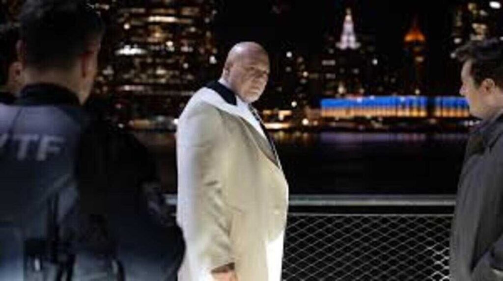 Cena noturna em um cais com a linha do horizonte de uma cidade iluminada ao fundo. No centro, o personagem Wilson Fisk, o Rei do Crime, um homem careca e imponente vestindo um paletó branco, olha fixamente para alguém fora de quadro. Ele está posicionado entre um agente da lei à esquerda e outro homem à direita, sugerindo um momento de confronto ou custódia.