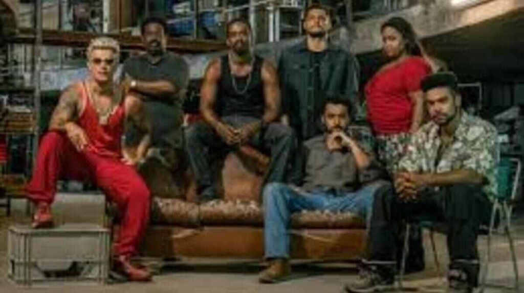 Foto em grupo do elenco da série Impuros em um cenário que remete a um galpão industrial. Vários homens e uma mulher posam com expressões sérias; alguns estão sentados em um sofá de couro desgastado e outros em pé ao redor. O homem à esquerda usa um conjunto vermelho vibrante e óculos escuros, enquanto os demais vestem roupas casuais em tons escuros e neutros.