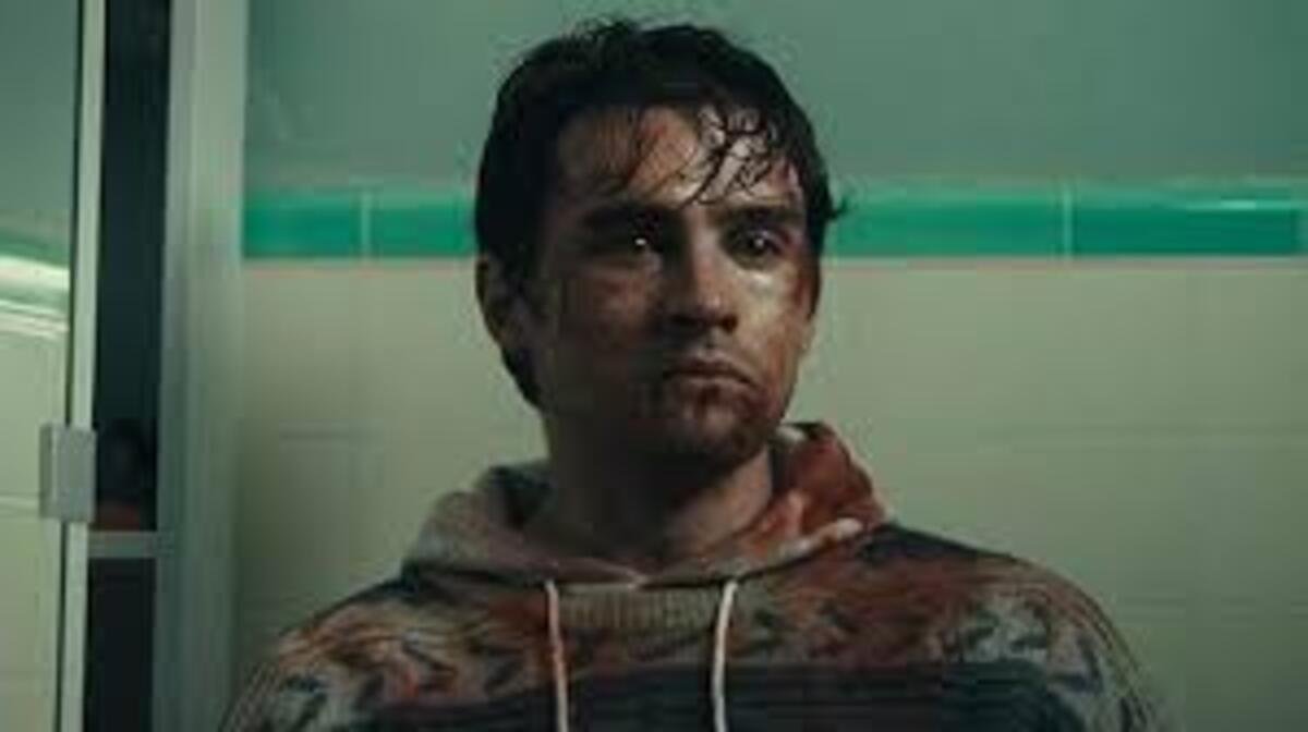 Um frame de filme mostrando o personagem de cabelos escuros e expressão tensa, após um confronto. Seus ferimentos e sangue no rosto indicam um evento violento recente. Ele veste um moletom com padrão tie-dye em tons terrosos. A cena tem uma atmosfera de drama e suspense.