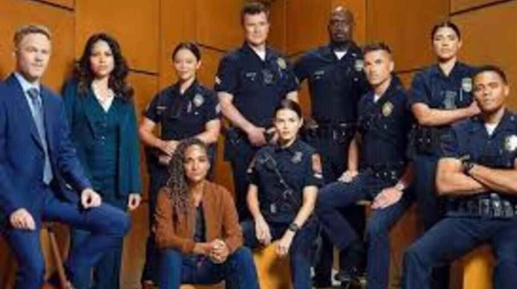 Foto promocional do elenco da série The Rookie reunido em um cenário com paredes de painéis de madeira clara. À esquerda, um homem loiro de terno azul e uma mulher de terninho azul-escuro aparecem posicionados. No centro e à direita, a maior parte do grupo veste uniformes escuros oficiais de patrulha da polícia, incluindo camisas de manga curta com distintivos e insígnias. Uma mulher de jaqueta marrom e calça jeans aparece sentada à frente, enquanto os demais integrantes, homens e mulheres, posam em pé ou apoiados em banquetas de madeira com expressões sérias e confiantes.