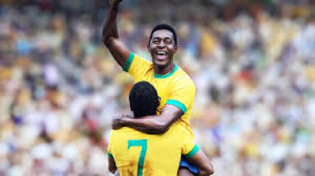 Crítica | Brasil 70 – A Saga do Tri: O gol de placa da Netflix que une futebol e ditadura militar 3 Reconstituição da histórica comemoração de Pelé e Jairzinho no gramado em cena da série Brasil Setenta A Saga do Tri. A imagem com forte apelo emocional e estético sobre a conquista do futebol brasileiro é perfeita para ilustrar resenhas de episódios, análises de produções documentais nacionais e listas de grandes momentos do esporte na televisão.