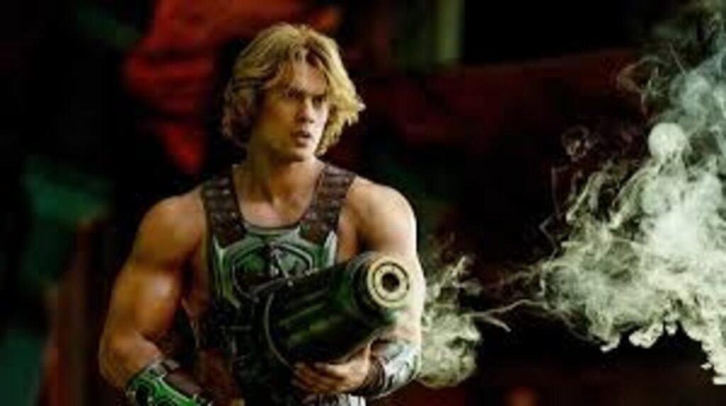Cena cinematográfica de ação em plano médio. O herói He-Man, interpretado por um ator loiro e musculoso com cabelos levemente ondulados, aparece posando de corpo inteiro da cintura para cima. Ele veste sua armadura tática de combate com tiras de couro marrom sobre os ombros e detalhes esverdeados no peito. O herói segura firmemente com as duas mãos uma grande arma futurista e cilíndrica de cor escura, que aponta para a direita enquanto libera uma densa cortina de fumaça esbranquiçada e cinzenta pelo cenário. O plano de fundo é escuro e sombrio, destacando os efeitos visuais do disparo.