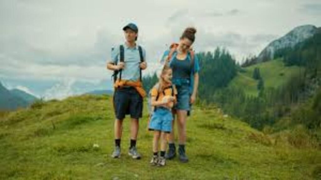 Fotografia colorida em plano médio de uma família praticando caminhada em uma região montanhosa. À esquerda, um homem jovem de boné escuro, camiseta polo azul-clara, bermuda escura e mochila de trilha nas costas observa a paisagem lateral, trazendo um casaco amarrado na cintura. À direita, uma mulher de cabelos presos em um coque e mochila nas costas caminha inclinando a cabeça com um sorriso carinhoso em direção a uma menina pequena que está entre o casal. A criança veste camiseta amarela, bermuda jeans clara e segura um bicho de pelúcia nos braços. Eles estão posicionados sobre uma colina gramada e verdejante, cercada por vales e montanhas com florestas sob um céu nublado.