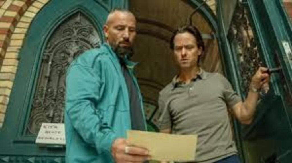 Cena cinematográfica em plano médio com ângulo de baixo para cima. Dois homens aparecem parados em frente a uma grande porta arqueada de cor esverdeada com detalhes ornamentais em ferro fundido. À esquerda, um homem musculoso, com barba escura e cabelos raspados nas laterais, veste um casaco esportivo verde-turquesa e segura um envelope ou papel amarelado em primeiro plano, olhando fixamente para ele. À direita, o segundo homem, de cabelos escuros e camisa polo cinza, olha para o mesmo papel com uma expressão de dúvida ou preocupação enquanto segura a maçaneta da porta aberta. A parede ao fundo revela tijolos à vista na entrada do edifício.