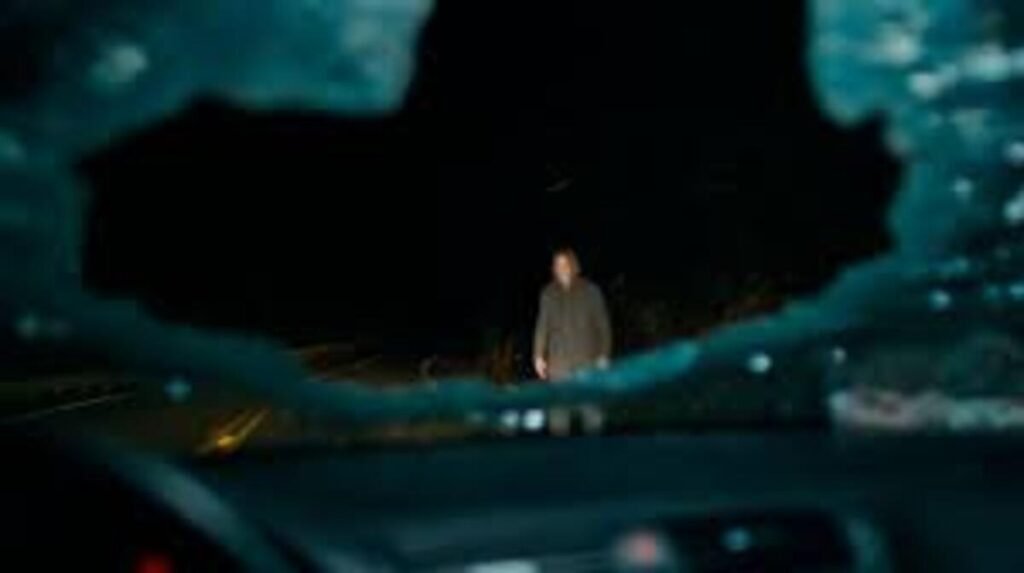 Ponto de vista de dentro de um veículo com o para-brisa estilhaçado mostrando uma figura misteriosa na estrada em cena do filme Passageiro do Mal. A imagem promocional de forte impacto visual e clima de suspense psicológico é excelente para ilustrar críticas de cinema de terror, análises de direção de fotografia e teorias sobre o enredo no blog.