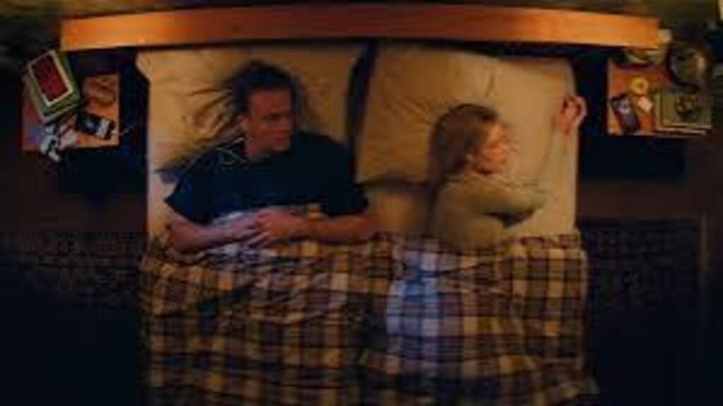 Casal distante deitado na cama em plano zenital durante cena de drama e mistério do filme Por Cima do Seu Cadáver. A imagem promocional focada na crise conjugal e no suspense psicológico dos protagonistas é excelente para ilustrar críticas de cinema, análises de relacionamento e resumos de enredos no blog.