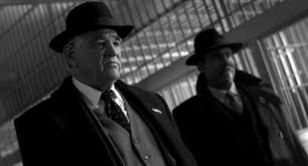 Crítica | Spider-Noir (1ª Temporada): Nicolas Cage brilha no espetacular suspense policial do Prime Video 5 Cena cinematográfica em preto e branco com enquadramento em plano médio e ângulo ligeiramente inclinado. Em primeiro plano, à esquerda, aparece um homem mais velho com expressão séria e compenetrada, vestindo um sobretudo escuro, terno com colete abotoado, camisa clara e gravata estampada, além de um chapéu de feltro clássico estilo fedora. Um pouco mais ao fundo e à direita, um segundo homem com bigode, vestindo trajes semelhantes com sobretudo e chapéu, acompanha-o com o olhar fixo na mesma direção. O cenário ao fundo apresenta linhas estruturais diagonais que lembram a arquitetura de uma grande estação, saguão ou grades de uma prisão.