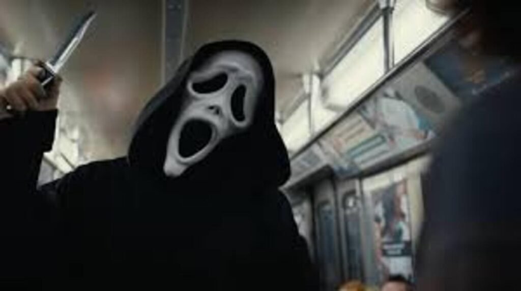 Crítica | Todo Mundo em Pânico 6: A nostalgia não salva esta crônica de um fracasso anunciado 2 Cena cinematográfica de suspense em plano médio com iluminação fria. O vilão Ghostface aparece em destaque no interior de um vagão de metrô em movimento, vestindo sua icônica túnica preta com capuz escuro. Sua máscara branca com expressão de pânico e boca aberta está levemente inclinada. Com a mão direita levantada na altura do ombro, ele segura firmemente uma faca metálica de lâmina afiada e brilhante, em posição de ataque. À direita, em primeiríssimo plano e de costas, aparece a silhueta desfocada de uma possível vítima vestindo roupa escura. O fundo do vagão exibe barras de metal para apoio e anúncios publicitários nas paredes laterais.