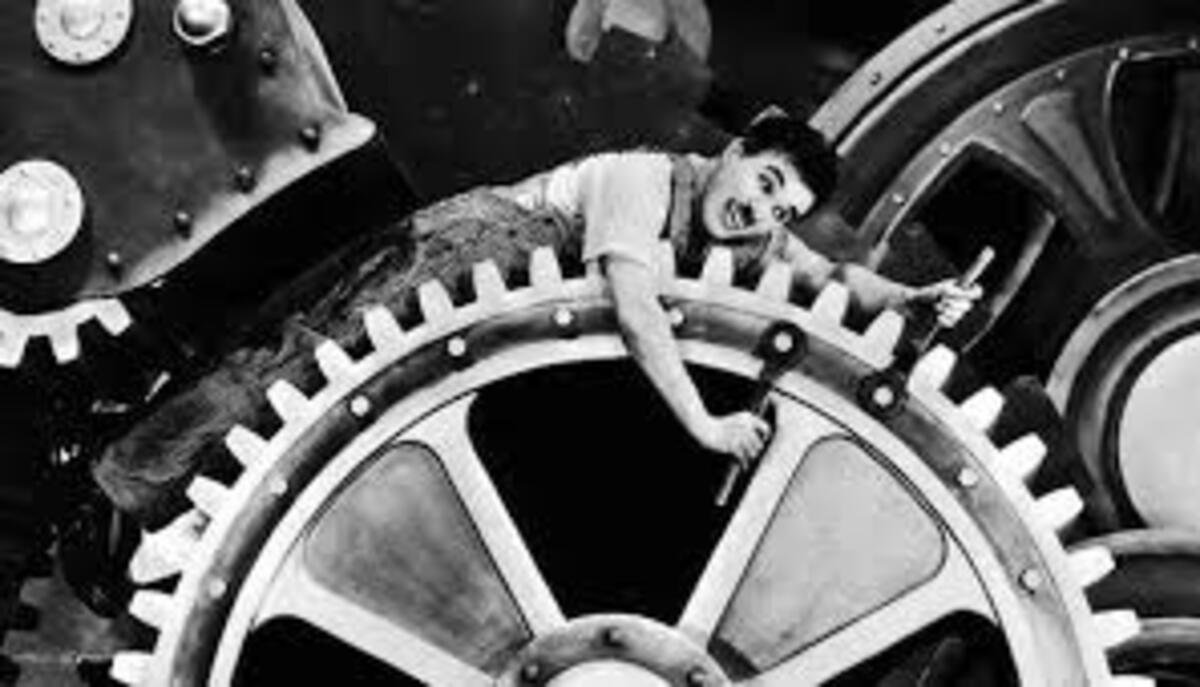 Cena cinematográfica icônica em preto e branco. O personagem Vagabundo, interpretado por Charles Chaplin, aparece deitado e preso de forma cômica entre os dentes de uma enorme engrenagem industrial de metal. Ele veste sua clássica calça larga com suspensórios e camiseta clara, segurando uma ferramenta em uma das mãos enquanto seu corpo acompanha a curvatura da grande peça redonda. Ao fundo, outras partes de maquinários pesados completam o cenário da fábrica.