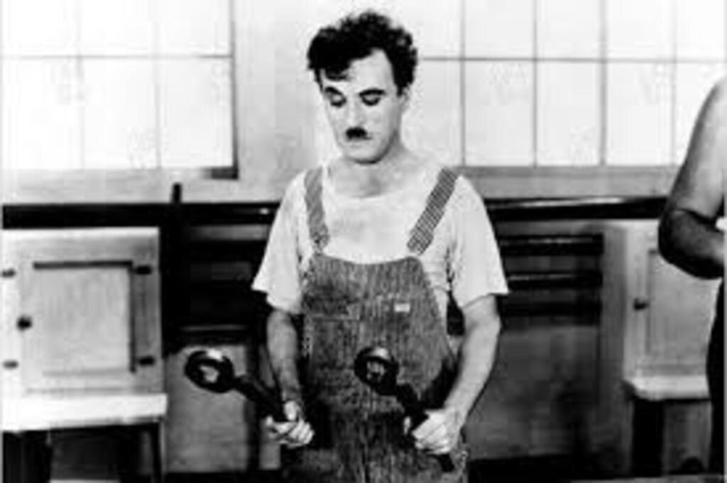 Tempos Modernos (1936): A Crítica de Charlie Chaplin à Grande Depressão e ao Fordismo 6 Charles Chaplin segurando ferramentas de fábrica em uma cena emblemática de Tempos Modernos. A imagem clássica em preto e branco que retrata a exaustão e a fixação do operário com seu trabalho repetitivo é ideal para ilustrar críticas de cinema, análises sobre a obra de Chaplin e listas de grandes momentos do cinema mudo no site.