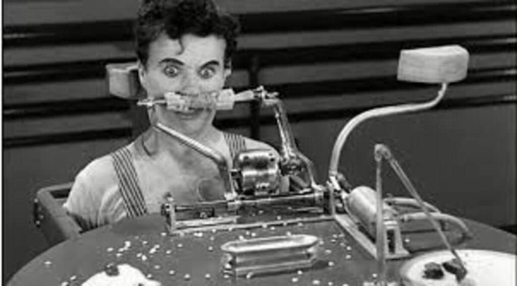 Tempos Modernos (1936): A Crítica de Charlie Chaplin à Grande Depressão e ao Fordismo 5 Cena clássica de cinema em preto e branco. O personagem de Charles Chaplin aparece preso a uma engenhosa máquina automática de alimentar disposta sobre uma mesa redonda. Ele veste seu macacão de operário com alças listradas e está com os olhos arregalados em uma expressão de completo espanto, choque e desconforto. Um braço mecânico do aparelho segura uma espiga de milho diretamente contra a sua boca. À frente, sobre a mesa salpicada de grãos, há pequenas bandejas de metal e outras partes articuladas do mecanismo, incluindo um braço curvo superior com uma esponja na ponta.