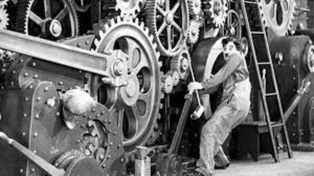 Tempos Modernos (1936): A Crítica de Charlie Chaplin à Grande Depressão e ao Fordismo 2 Cena cinematográfica em preto e branco mostrando o personagem de Charles Chaplin trabalhando em uma fábrica complexa e repleta de maquinários pesados. À direita, Chaplin aparece em pé, vestindo um macacão de operário com suspensórios e ostentando seu clássico bigode. Ele está inclinado para trás, segurando com as duas mãos uma longa alavanca de metal conectada à máquina. À esquerda, o cenário é dominado por um enorme sistema interligado de grandes rodas dentadas e engrenagens industriais. Uma escada de mão comprida está apoiada na lateral direita da máquina.