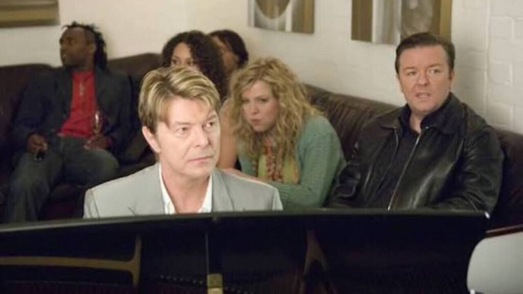 Famosos na TV: Participações especiais em séries que você pode ter deixado passar 7 O astro da música David Bowie tocando piano ao lado de Ricky Gervais em cena memorável de famosa série de comédia britânica na televisão. A imagem capturando uma das participações especiais mais icônicas e celebradas da cultura pop é perfeita para ilustrar listas de curiosidades, artigos sobre bastidores e análises de episódios clássicos no site.