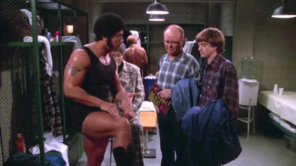 Famosos na TV: Participações especiais em séries que você pode ter deixado passar 5 Participação especial icônica do ator Dwayne Johnson interpretando um lutador com visual retrô em cena de famosa série de comédia de televisão. A imagem de bastidores e divulgação destacando momentos nostálgicos da cultura pop é ideal para ilustrar críticas de episódios, listas de participações de astros de Hollywood e artigos de entretenimento no site.