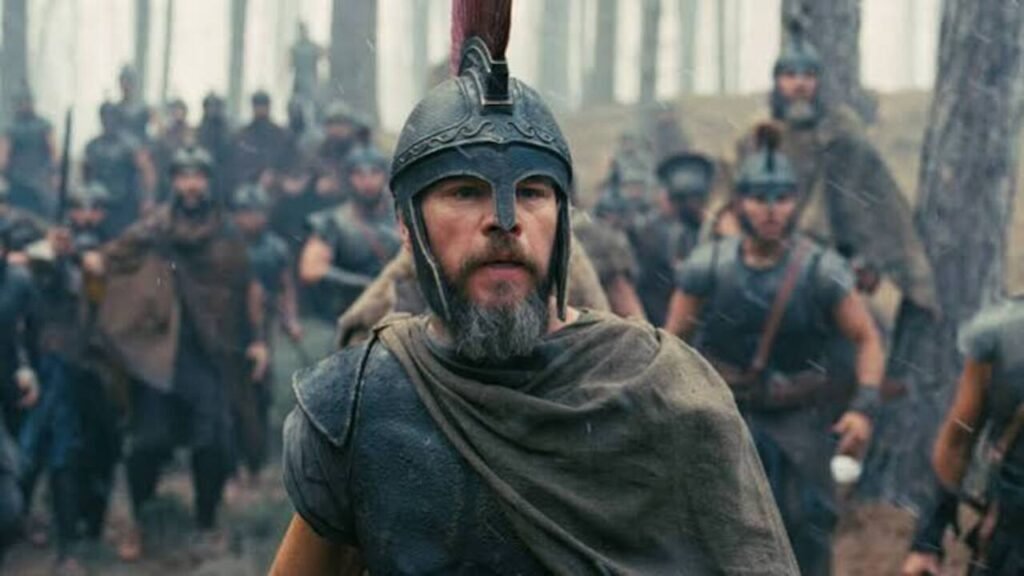 Em primeiro plano, um homem com barba cinzenta vestindo armadura e capacete de estilo grego antigo com pluma vermelha avança com expressão séria. Ao fundo, uma tropa de soldados equipados marcha através de uma floresta.