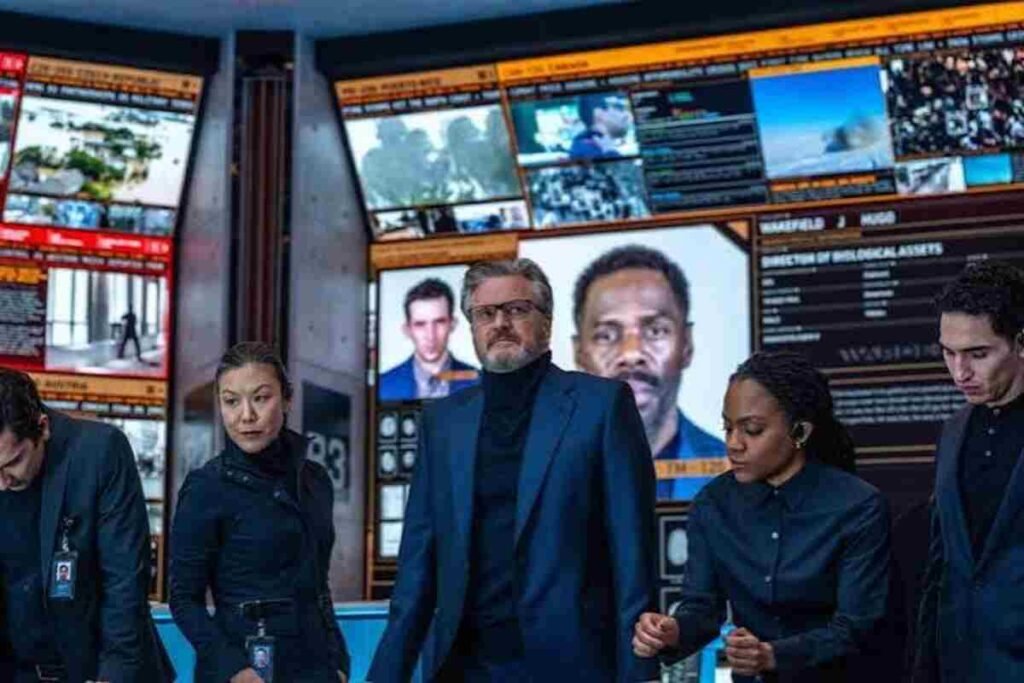 Crítica | Dia D (2026): O novo filme de ficção científica de Steven Spielberg decepciona? 2 Colin Firth de paletó azul e óculos lidera equipe de agentes de preto em frente a grandes telas de monitoramento em sala de controle de alta tecnologia.