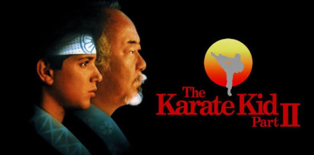 Pôster do filme Karate Kid Parte II mostrando o perfil de Daniel LaRusso e Senhor Miyagi ao lado do logotipo do filme com um sol nascente.